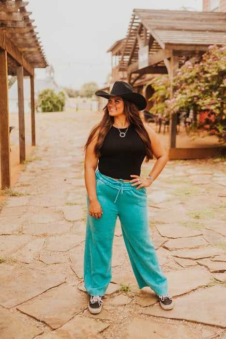 Woman wearing a black cowboy hat, black top, and turquoise pants standing on a stone path.