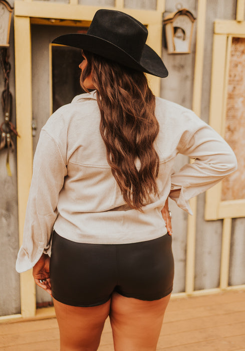 Person wearing a black cowboy hat and light-colored shirt with black shorts, standing in front of wooden structures.