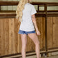 Woman standing in a stable wearing a white t-shirt and denim shorts.