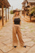 Woman in black top and beige pants standing on a stone path with wooden structures and plants in the background.