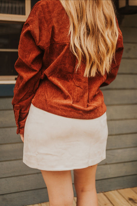 Person wearing a rust-colored corduroy jacket and white skirt on a wooden deck.