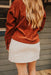 Person wearing a rust-colored corduroy jacket and white skirt on a wooden deck.