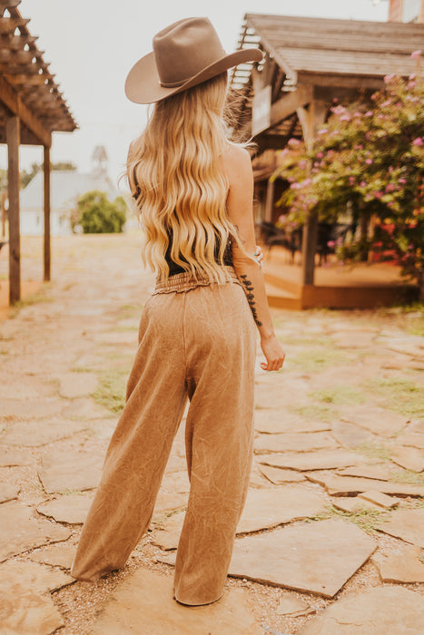 Woman in a brown hat and wide-leg pants walking on a stone path with a garden in the background