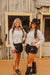 Two women in white shirts and black shorts standing in front of a wooden building.