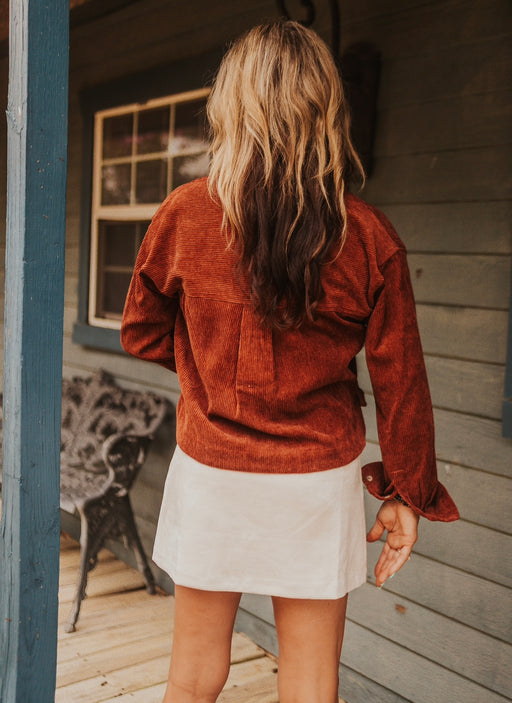 Person wearing a rust-colored jacket and white skirt standing on a wooden deck.