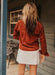 Person wearing a rust-colored jacket and white skirt standing on a wooden deck.