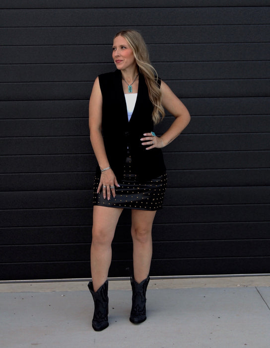 Woman wearing a black vest over a white top and black polka dot skirt against a black background