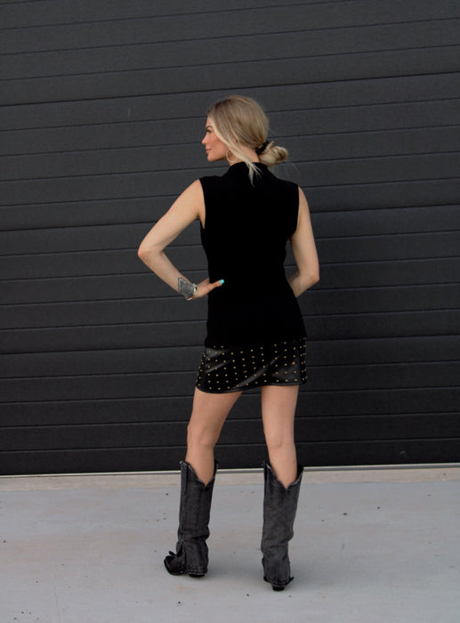 Woman wearing a black sleeveless top and skirt standing against a black wall.