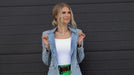 Woman wearing a denim jacket, white top, green shorts, and denim boots against a black wall.