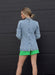 Woman wearing a denim jacket, green shorts, and blue cowboy boots against a black wall.