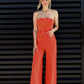 Woman wearing a bright orange strapless jumpsuit against a black slatted wall.