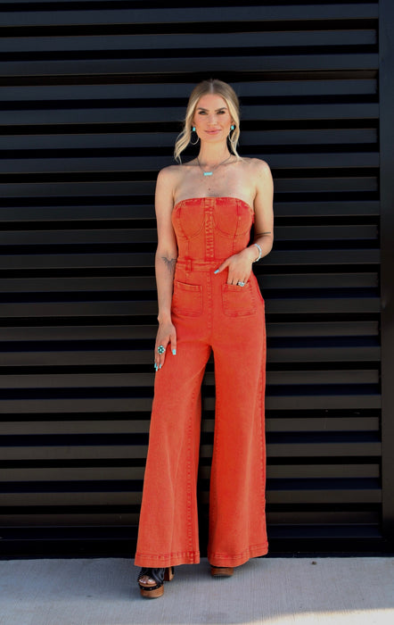 Woman wearing a bright orange strapless jumpsuit against a black slatted wall.