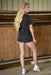 Woman standing in a stable wearing a dark denim shirt and shorts.