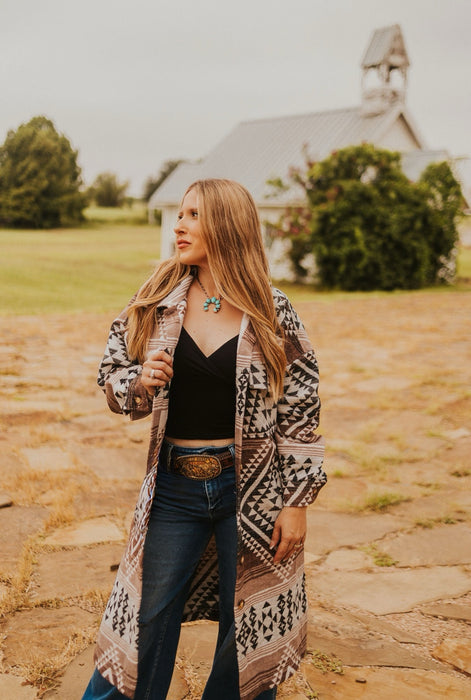 Woman standing in a field wearing a patterned coat and black top, with a church in the background.