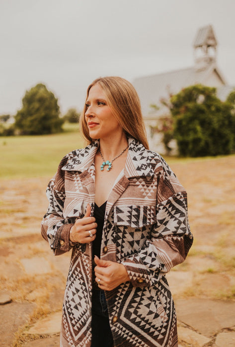 Woman wearing a patterned jacket outdoors with a building and greenery in the background