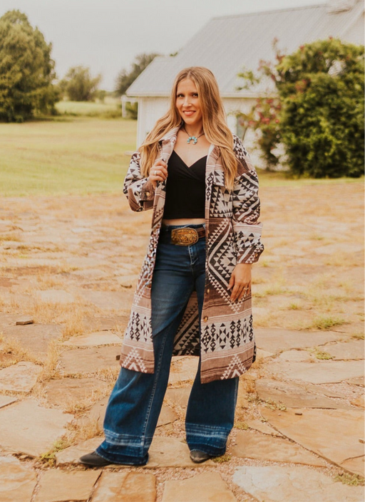 Woman in a patterned coat and jeans standing on a stone path outdoors.
