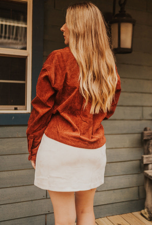Woman wearing a rust-colored jacket and white skirt standing on a wooden deck.