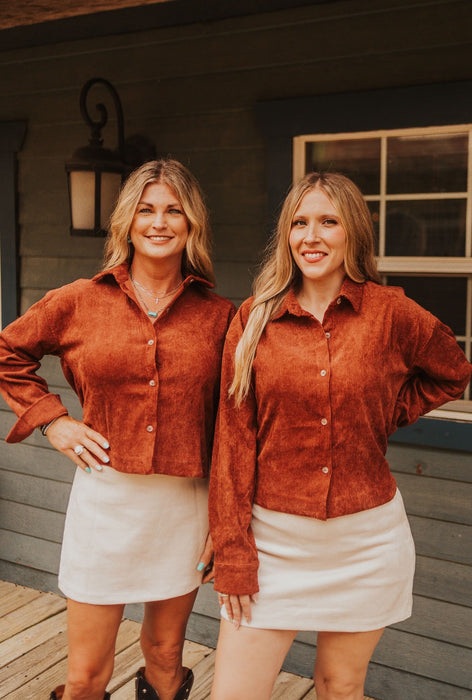 Two women wearing matching rust-colored button-up shirts and white skirts standing outdoors.