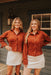 Two women wearing matching rust-colored button-up shirts and white skirts standing outdoors.