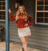 Woman in a red jacket and white skirt standing on a wooden porch.