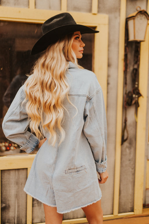Woman wearing a denim jacket and black hat in front of a rustic wooden wall.