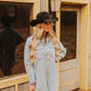 Woman wearing a denim dress and black cowboy hat standing in front of a wooden building.