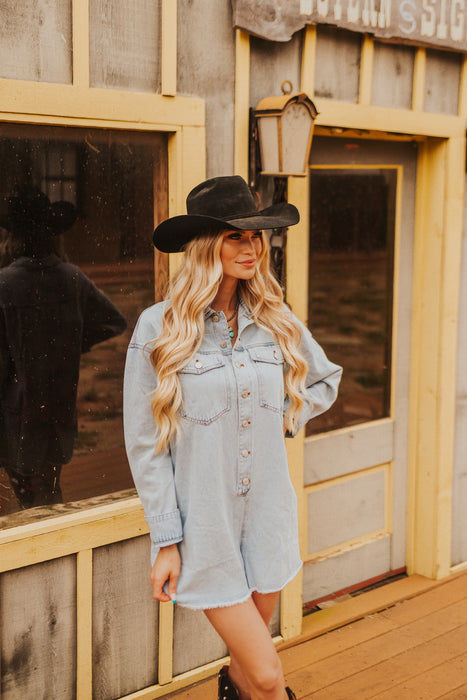 Woman wearing a denim dress and black cowboy hat standing in front of a wooden building.