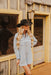 Woman wearing a denim dress and black cowboy hat standing in front of a wooden building.