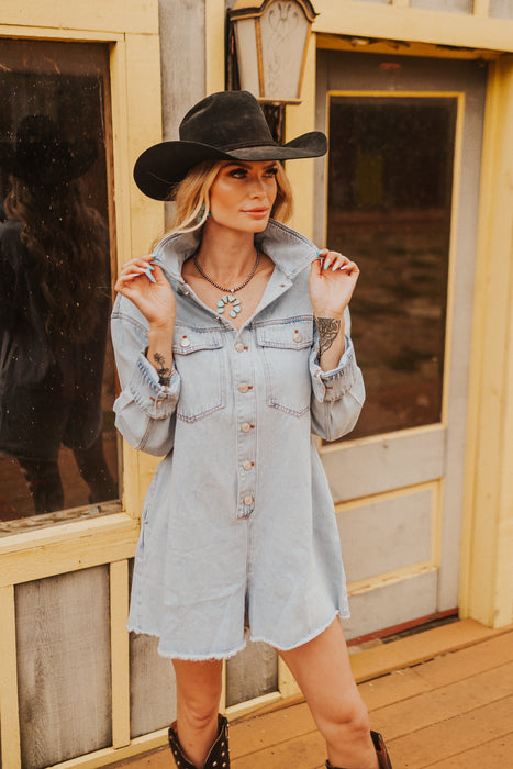 Woman wearing a denim dress and cowboy hat in front of a wooden building.