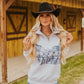 Woman wearing a white hoodie with a graphic design and a black cowboy hat in front of a wooden building.