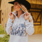 Woman wearing a white hoodie with a graphic design, black hat, and blue jeans in front of a wooden building.
