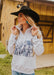 Woman wearing a white hoodie with a graphic design, black hat, and blue jeans in front of a wooden building.