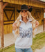 Woman wearing a white hoodie with a graphic design and a black cowboy hat in front of a wooden building.