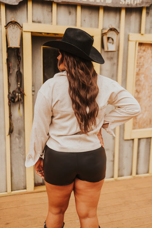 Person wearing a black cowboy hat and light shirt standing in front of a wooden building.