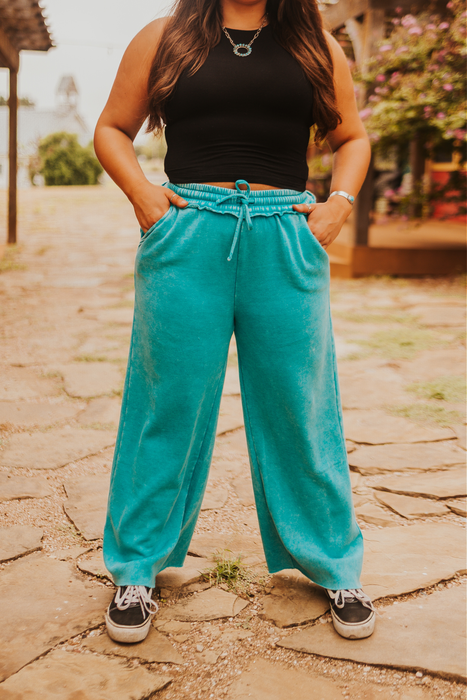 Person wearing turquoise pants and a black top standing on a stone path with greenery in the background