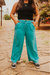 Person wearing turquoise pants and a black top standing on a stone path with greenery in the background