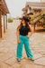 Woman wearing a black cowboy hat, black top, and turquoise pants standing on a stone path.