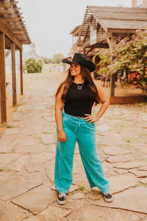 Woman wearing a black cowboy hat, black top, and turquoise pants standing on a stone path.