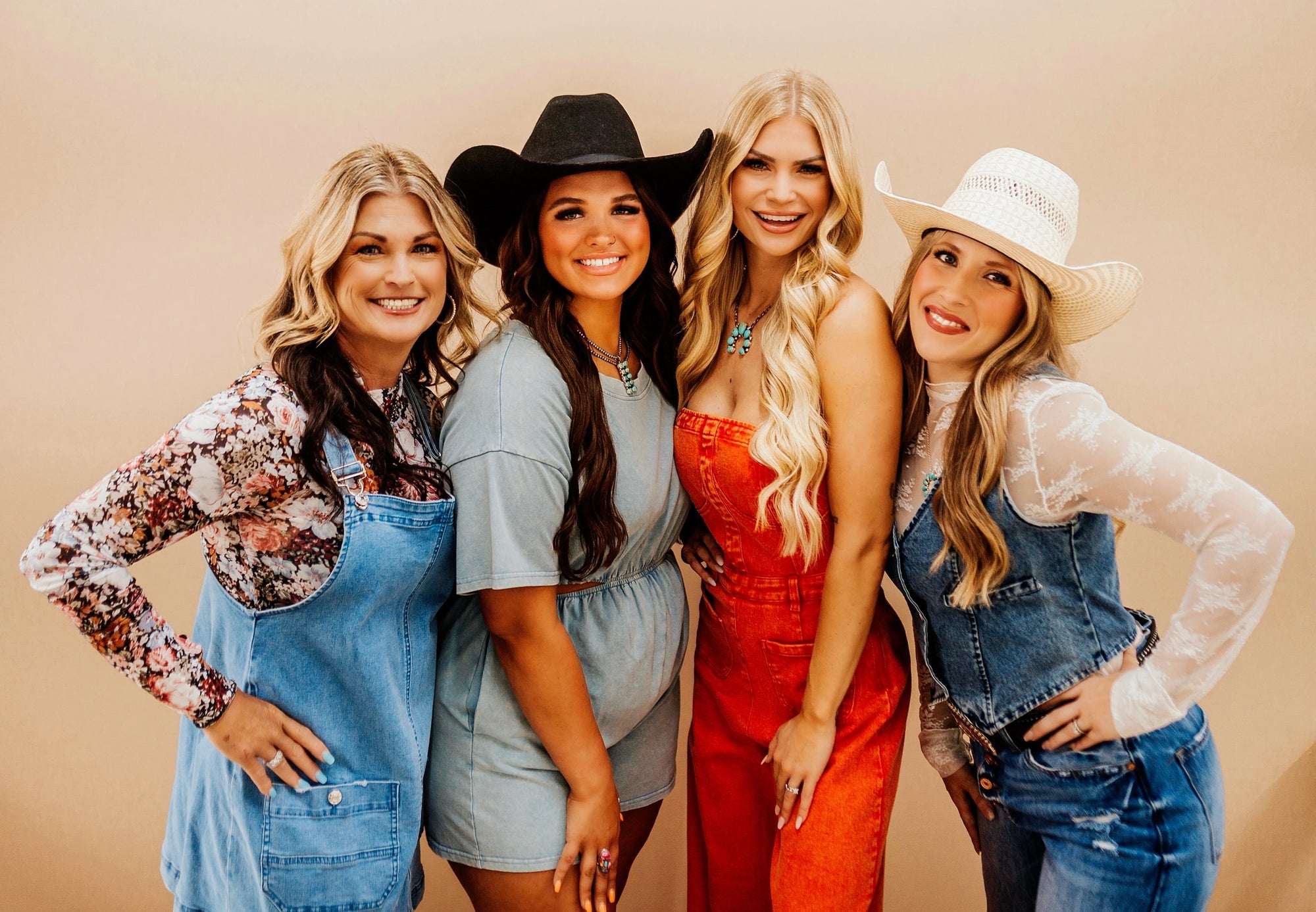 Four women posing together against a beige background, wearing cowboy hats and casual outfits.