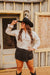 Woman wearing a white shirt and black shorts with a cowboy hat, standing in front of a rustic wooden building.