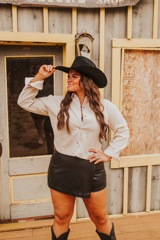 Woman wearing a white shirt and black shorts with a cowboy hat, standing in front of a rustic wooden building.