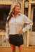 Woman wearing a beige button-up shirt and black skirt standing in front of a rustic building.