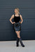 Woman wearing a black top and sequin skirt with black knee-high boots against a dark wall.