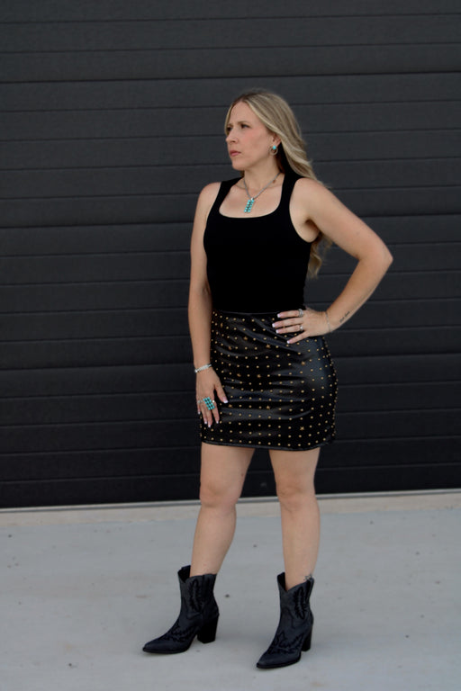 Woman wearing a black tank top and studded black skirt with black boots against a black background