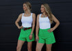 Two women wearing matching outfits with white tops and green shorts against a black background.