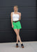 Woman wearing a white tank top, green shorts, and black shoes against a black background