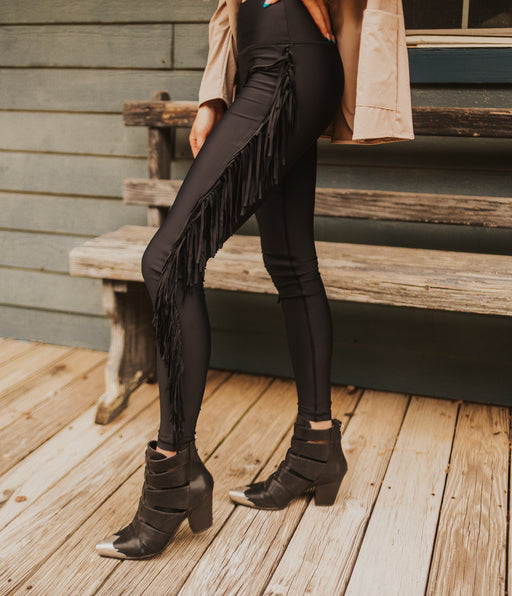 Person wearing black fringed leggings and boots on a wooden deck.