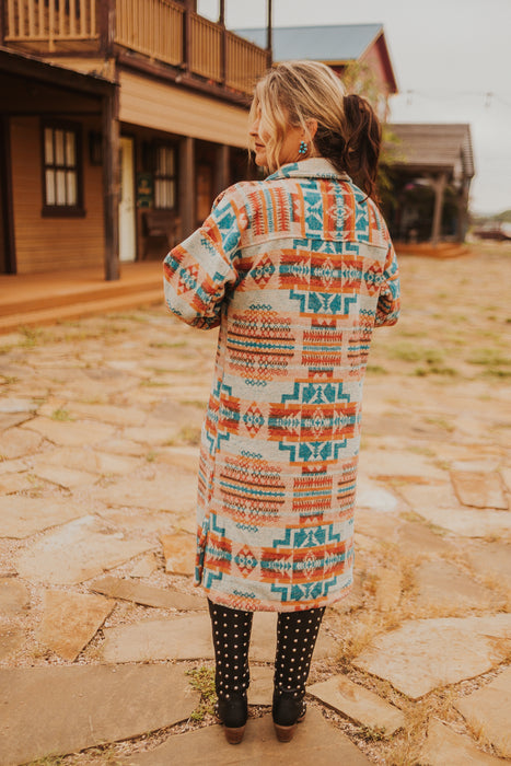 Woman wearing a colorful patterned coat standing on a stone path with wooden buildings in the background.