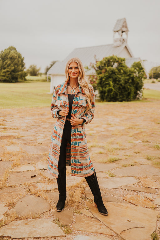 Woman in a colorful coat standing on a stone path with a house and trees in the background