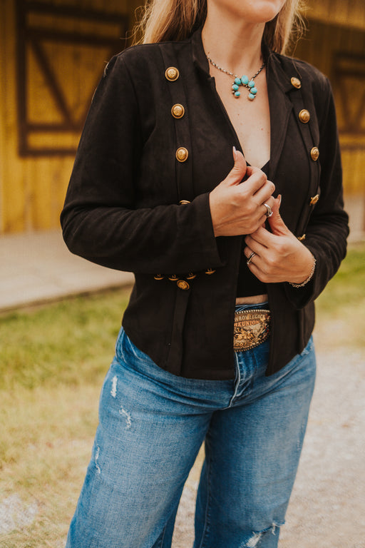 Person wearing a black jacket with gold buttons and blue jeans outdoors.